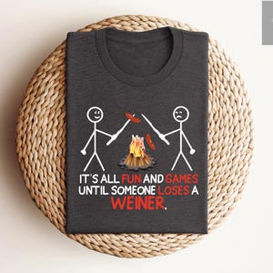 May include: Dark gray t-shirt with a humorous graphic. The design features stick figures roasting hot dogs over a campfire, with the text "IT'S ALL FUN AND GAMES UNTIL SOMEONE LOSES A WEINER." The shirt is displayed on a woven, circular placemat.