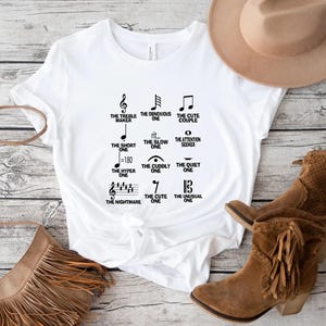 May include: White t-shirt with black musical note graphics and text. The shirt features various musical note designs with phrases like "The Treble Maker," "The Cute Couple," and "The Nightmare." A brown hat and boots are in the background.