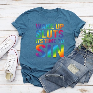 May include: Teal t-shirt with the text "WAKE UP SLUTS IT'S TIME TO SIN" in a rainbow gradient. The shirt is paired with blue denim jeans and white canvas sneakers. A small green plant is also in the image.