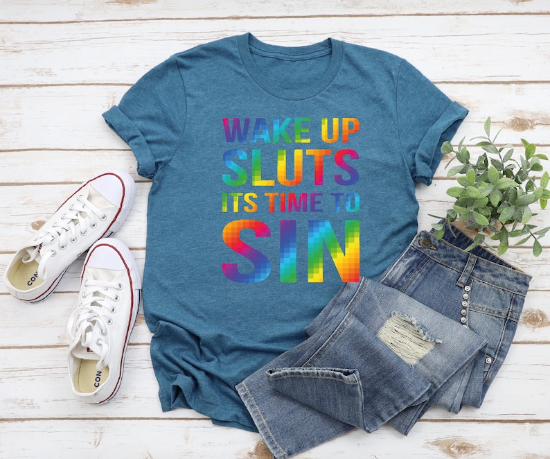 May include: Teal t-shirt with the text "WAKE UP SLUTS IT'S TIME TO SIN" in a rainbow gradient. The shirt is paired with blue denim jeans and white canvas sneakers. A small green plant is also in the image.