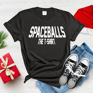 May include: Black t-shirt with white text that reads "Spaceballs The T-Shirt". The t-shirt is laid out on a white wooden surface with a pair of blue jeans, a red Santa hat, and a red gift box.