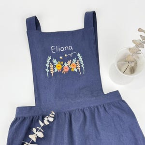 May include: A denim blue apron featuring floral embroidery and the name "Eliana". The apron has two front pockets and adjustable straps. The apron is made of a soft, durable fabric. The apron is suitable for cooking or crafting.