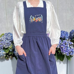 May include: A navy blue pinafore dress with a floral embroidery design on the front. The dress has two pockets on the front and a gathered skirt.