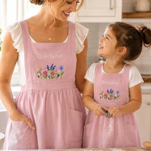 Peut inclure: Tabliers rose poudré assortis avec les prénoms Oaklynn et Brookie brodés en blanc au-dessus de motifs floraux colorés. Chaque tablier présente une encolure carrée, de larges bretelles et deux poches avant, portés sur des t-shirts blancs.