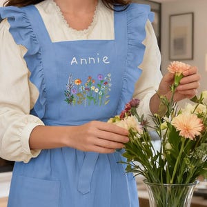 May include: A pale blue apron with frilled edges and a front pocket. The apron is embroidered with the name "Annie" and a floral design. A person is arranging flowers in a glass vase. The apron is worn over a white blouse.