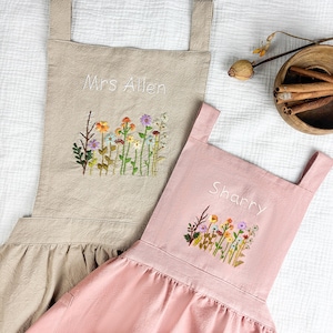 May include: Two aprons, one beige and one pink, each featuring embroidered wildflower designs and personalised names. The beige apron is embroidered with "Mrs Allen" and the pink apron with "Sharry" above colourful floral stems.