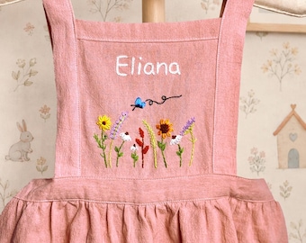 May include: A dusty pink pinafore dress featuring white embroidered text that reads Eliana, positioned above a colorful floral embroidery design with a small blue butterfly. The garment has wide shoulder straps and a gathered skirt with pockets.
