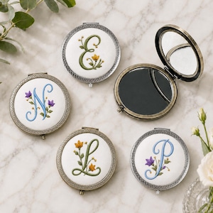 May include: Four round compact mirrors with embroidered white fabric covers featuring the letters E, N, L, and P in cursive script, accented with small floral designs. One mirror is shown open, revealing a reflective surface and a metallic frame.