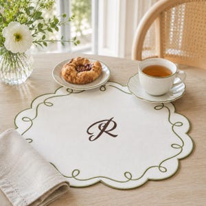 May include: A round white placemat featuring a dark green scalloped embroidered edge and a central dark brown embroidered letter R monogram. The placemat is placed on a wooden table with a white flower vase, a pastry on a plate, and a tea cup.