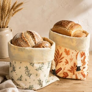 May include: Two fabric bread bags with roll-top closures and black plastic buckles. One bag features a green botanical leaf print on a cream background, while the other displays an orange and brown autumn-themed pattern of wheat and leaves.
