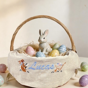 May include: An Easter basket with a woven handle, filled with decorative eggs and straw. A ceramic bunny sits inside. The basket's fabric is embroidered with the name "Lucas", a bunny, and a wheelbarrow.