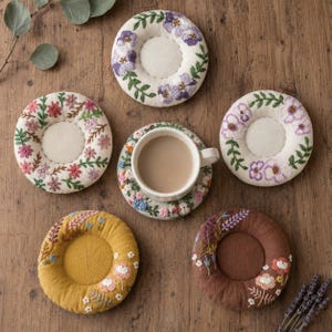 May include: Five circular, padded coasters featuring intricate floral embroidery in various colors, including purple, pink, yellow, and brown. One coaster supports a white ceramic mug filled with a light brown beverage, while the others are empty.