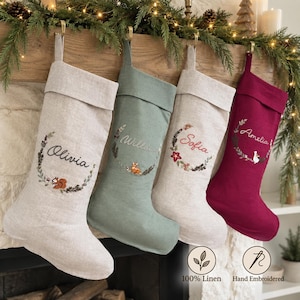 May include: Four hanging Christmas stockings in beige, sage green, light grey, and deep red linen. Each stocking features a folded cuff, a circular embroidered wreath design, and a personalised name: Olivia, Willow, Sofia, and Amelia.
