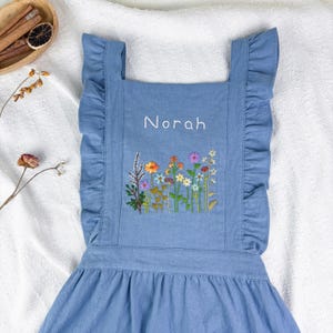 May include: A light blue apron with ruffled edges and straps. The apron features an embroidered floral design with the name "Norah" in white script. The flowers are in various colors, including orange, yellow, pink, and purple.