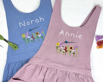 May include: Two sleeveless aprons in blue and mauve, each featuring white embroidered names Norah and Annie. Below the names, colorful floral embroidery depicts wildflowers and a small blue butterfly. The aprons have a gathered waist and pockets.