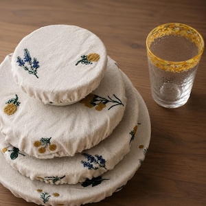 May include: A stack of four round, off-white fabric bowl covers featuring embroidered botanical motifs in yellow, blue, and green. Each cover has an elasticized edge. Beside the stack is a clear glass with a textured surface and a yellow rim.