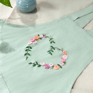 May include: A light green fabric with a heart-shaped floral embroidery design. The embroidery features pink, peach, and white flowers with green leaves.