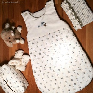 May include: A white baby sleeping bag with a blue and brown floral pattern. The sleeping bag has a white trim and a small embroidered flower design on the chest. The sleeping bag is on a wooden surface with a stuffed animal, a pair of booties, and a small quilted bag.