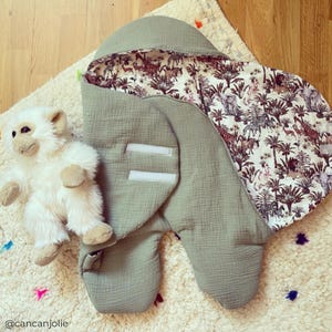 May include: A green muslin swaddle with a white and brown jungle animal print lining. The swaddle has a hood and velcro closures. A white stuffed monkey is lying next to the swaddle.