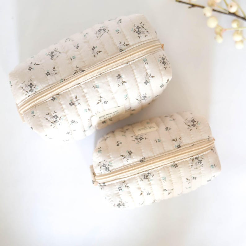 May include: Two quilted zippered pouches with a floral print. The pouches are beige with a small floral pattern in shades of gray and blue.