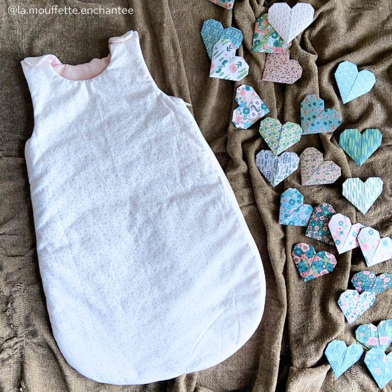 May include: A white sleeping bag with a pink lining and a small floral pattern. The sleeping bag is lying on a brown fabric background with a collection of colorful origami hearts.