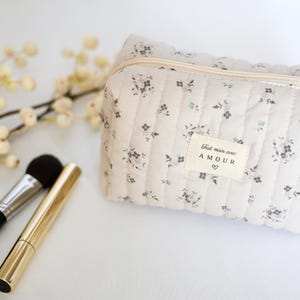 May include: A beige quilted makeup bag with a floral print and a zipper closure. The bag has a label that says "Fait main avec AMOUR" with a heart symbol.