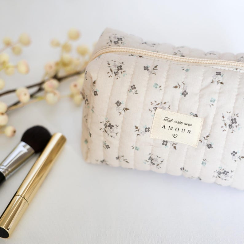 May include: A beige quilted makeup bag with a floral print and a zipper closure. The bag has a label that says "Fait main avec AMOUR" with a heart symbol.