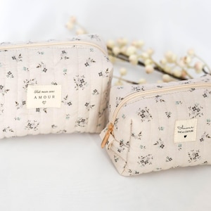 May include: Two beige quilted makeup bags with floral patterns. The larger bag has a label that reads "Fait main avec AMOUR". The smaller bag has a label that reads "Amour Tie & Couture".