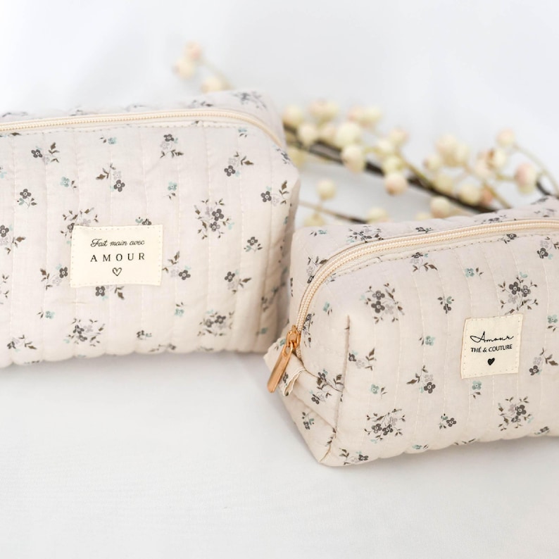 May include: Two beige quilted makeup bags with floral patterns. The larger bag has a label that reads "Fait main avec AMOUR". The smaller bag has a label that reads "Amour Tie & Couture".