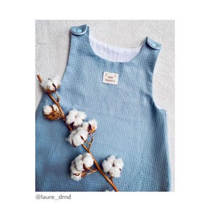 May include: A light blue baby onesie with a white trim and a small brown label that says "Petit Chat" on it. The onesie is photographed on a white background with a cotton branch.