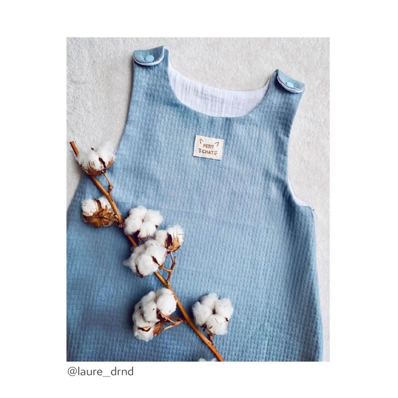May include: A light blue baby onesie with a white trim and a small brown label that says "Petit Chat" on it. The onesie is photographed on a white background with a cotton branch.