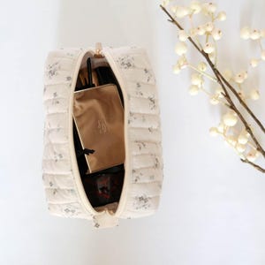 May include: A white quilted makeup bag with a floral print. The bag is open and contains a gold makeup case, a black makeup brush, and other makeup items.