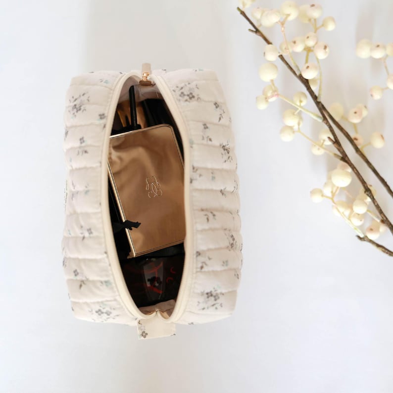 May include: A white quilted makeup bag with a floral print. The bag is open and contains a gold makeup case, a black makeup brush, and other makeup items.