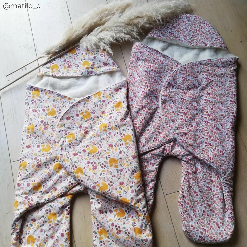 May include: Two baby swaddles with floral patterns. One swaddle is white with yellow foxes and the other is pink with red flowers. Both swaddles have a white hood.