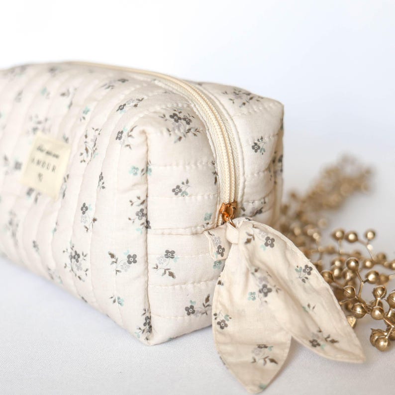 May include: A white quilted makeup bag with a floral pattern and a gold zipper. The bag has a small fabric loop attached to the zipper.