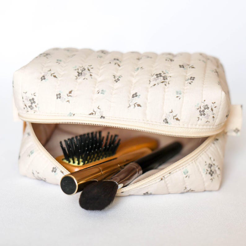 May include: A white quilted makeup bag with a floral print is open, revealing a black hairbrush, a wooden brush, and two makeup brushes.