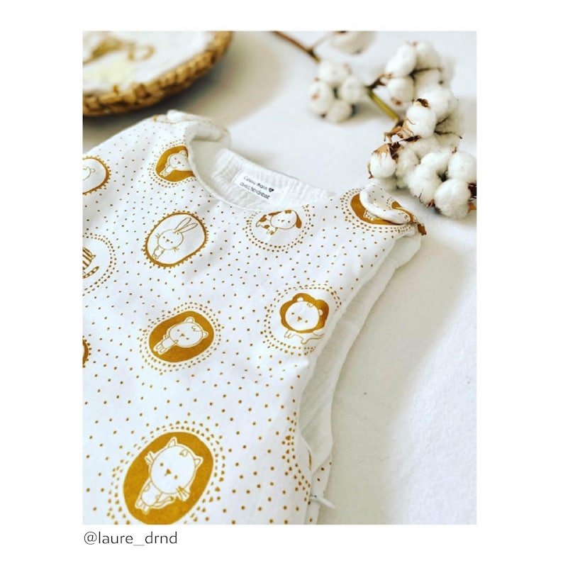 May include: White sleeveless sleep sack with a gold and white pattern of animals in circles. The pattern includes a lion, a rabbit, a cat, and a giraffe. The sleep sack has a zipper on the side.