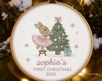 May include: Embroidered Christmas ornament featuring a bunny in a pink tutu, a Christmas tree, and the text "Sophia's First Christmas 2025." The ornament is set in a wooden hoop and hangs from a red ribbon, with a festive background.
