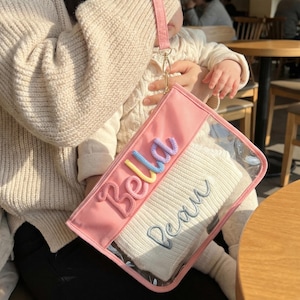 May include: A pink clutch with a clear front panel, featuring the embroidered names "Bella" and "Beau" in colorful and blue thread. The clutch has a pink wrist strap and contains a white item.
