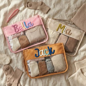Könnte beinhalten: Drei personalisierte Reisetaschen in Rosa, Beige und Orange, jede mit einem Namen in bunten Buchstaben bestickt: "Bella", "Mia" und "Jack". Die Taschen sind mit Socken und einem kleinen Pullover gefüllt. Zusätzliche Socken und Pullover sind verstreut.