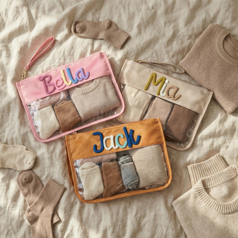 May include: Three personalized travel pouches in pink, beige, and orange, each with a name embroidered in colorful letters: "Bella", "Mia", and "Jack". The pouches are filled with socks and a small sweater. Additional socks and sweaters are scattered around.