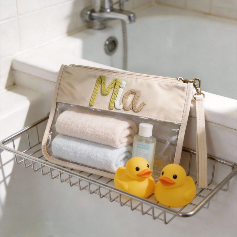 May include: A beige cosmetic bag with the name "Mia" in embroidered letters, containing folded towels and a small bottle. Two yellow rubber duck toys sit in front of the bag on a metal shelf.
