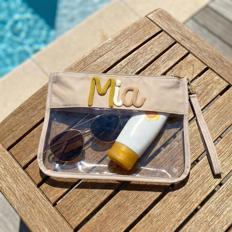 May include: A beige zippered pouch with a clear front panel, containing sunglasses and sunscreen. The name "Mia" is embroidered in yellow and tan thread on the top. The pouch has a wrist strap and is resting on a wooden surface.