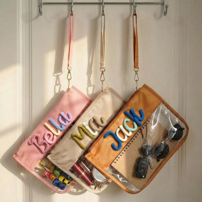 May include: Four personalized hanging pouches in pink, beige, and orange, each with a name embroidered in colorful yarn: Bella, Mia, and Jack. The pouches have clear front pockets and contain items like toys, makeup, and keys.