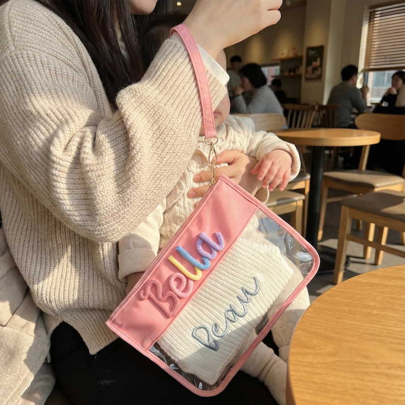 May include: A pink clutch with a clear front panel, featuring the embroidered names "Bella" and "Beau" in colorful and blue thread. The clutch has a pink wrist strap and contains a white item.
