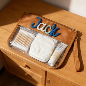 May include: A brown leather baby travel pouch with a clear front, containing baby essentials. The name "Jack" is embroidered on the pouch in blue and white yarn. Inside are cotton swabs, a diaper, and baby bum cream.