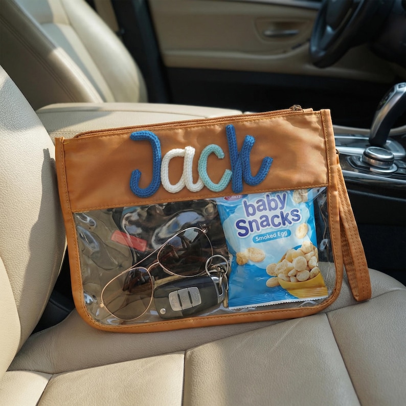 May include: A tan zippered pouch with a clear front panel, containing sunglasses, car keys, and a bag of baby snacks. The name "Jack" is embroidered in blue, white, and light blue yarn on the pouch.