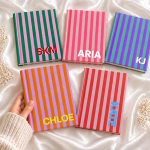 May include: Five colorful notebooks with vertical stripes in various colors, including green, pink, orange, red, and blue. Each notebook has a different name or initials in a contrasting color. The notebooks are arranged on a white satin surface.