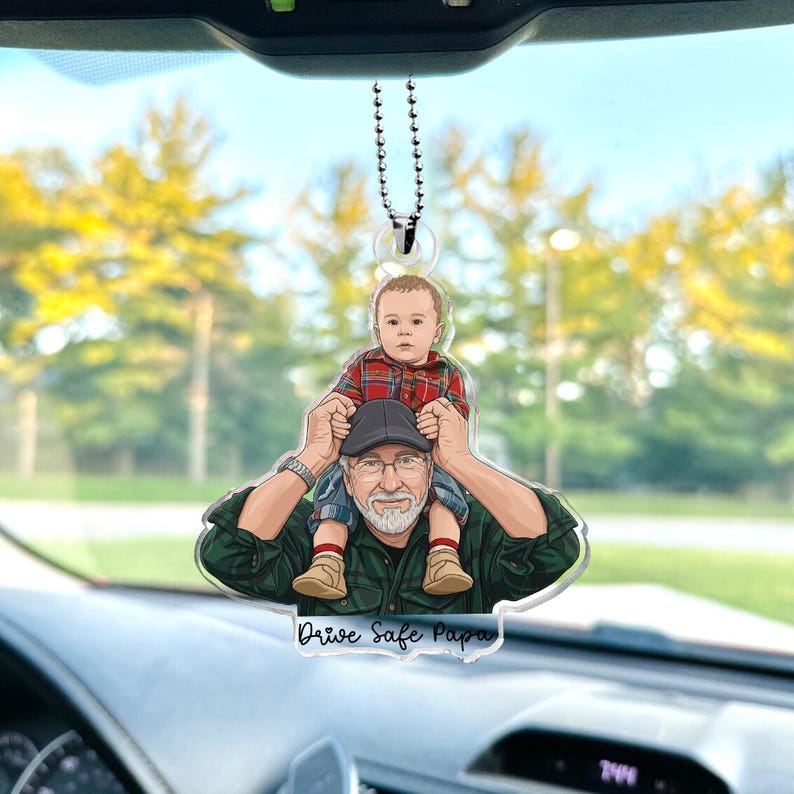May include: A car hanging ornament featuring a cartoon illustration of a man with a child on his shoulders. The child wears a red plaid shirt. The ornament has the text "Drive Safe Papa".