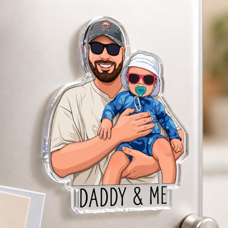 May include: Clear acrylic magnet featuring a cartoon illustration of a man holding a baby. Both are wearing sunglasses. The baby has a pacifier. The text "DADDY & ME" is at the bottom. The magnet is attached to a metallic surface.